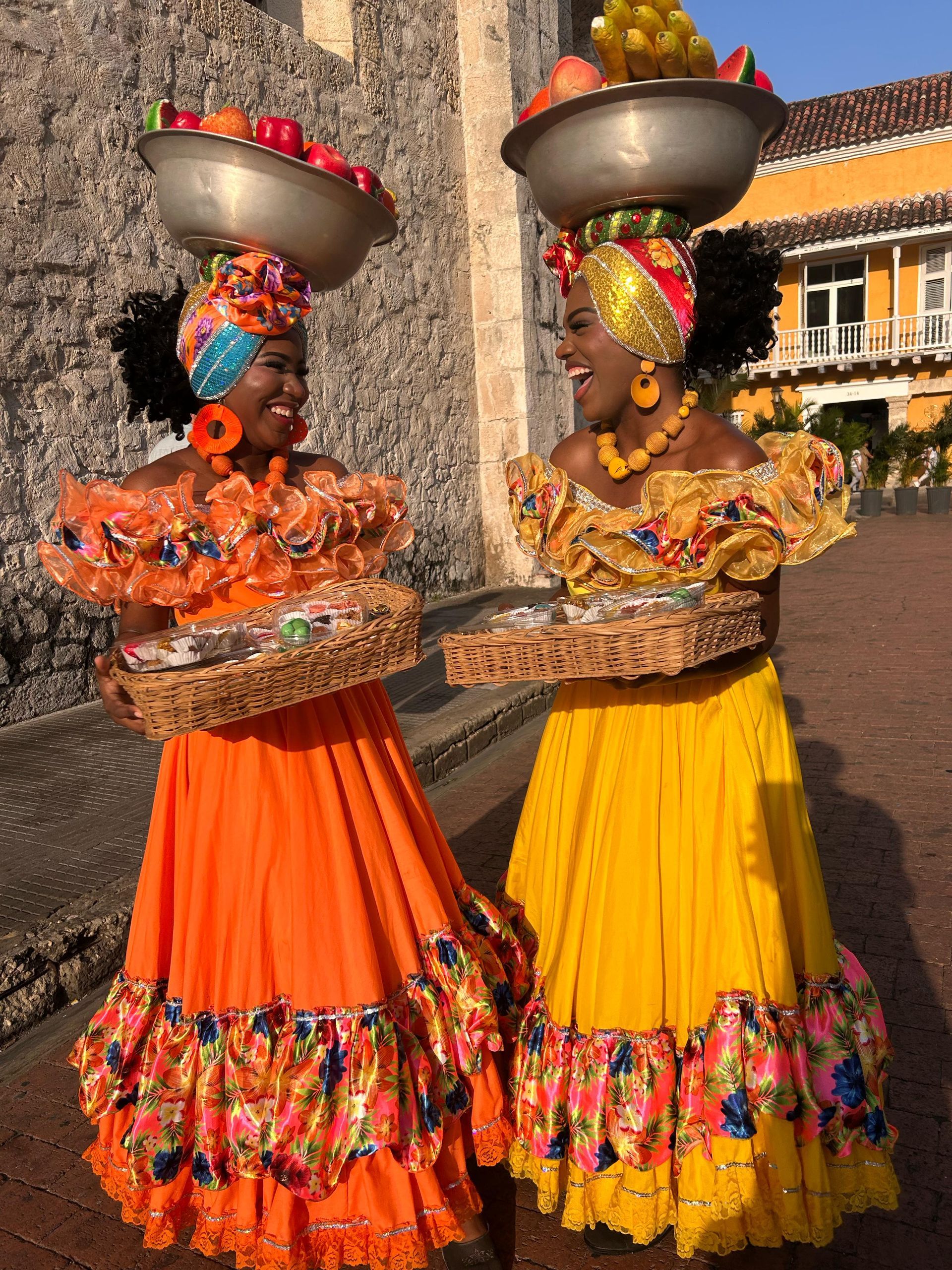 Dos mujeres sonrientes con coloridos vestidos tradicionales colombianos balancean fruteros sobre sus cabezas.