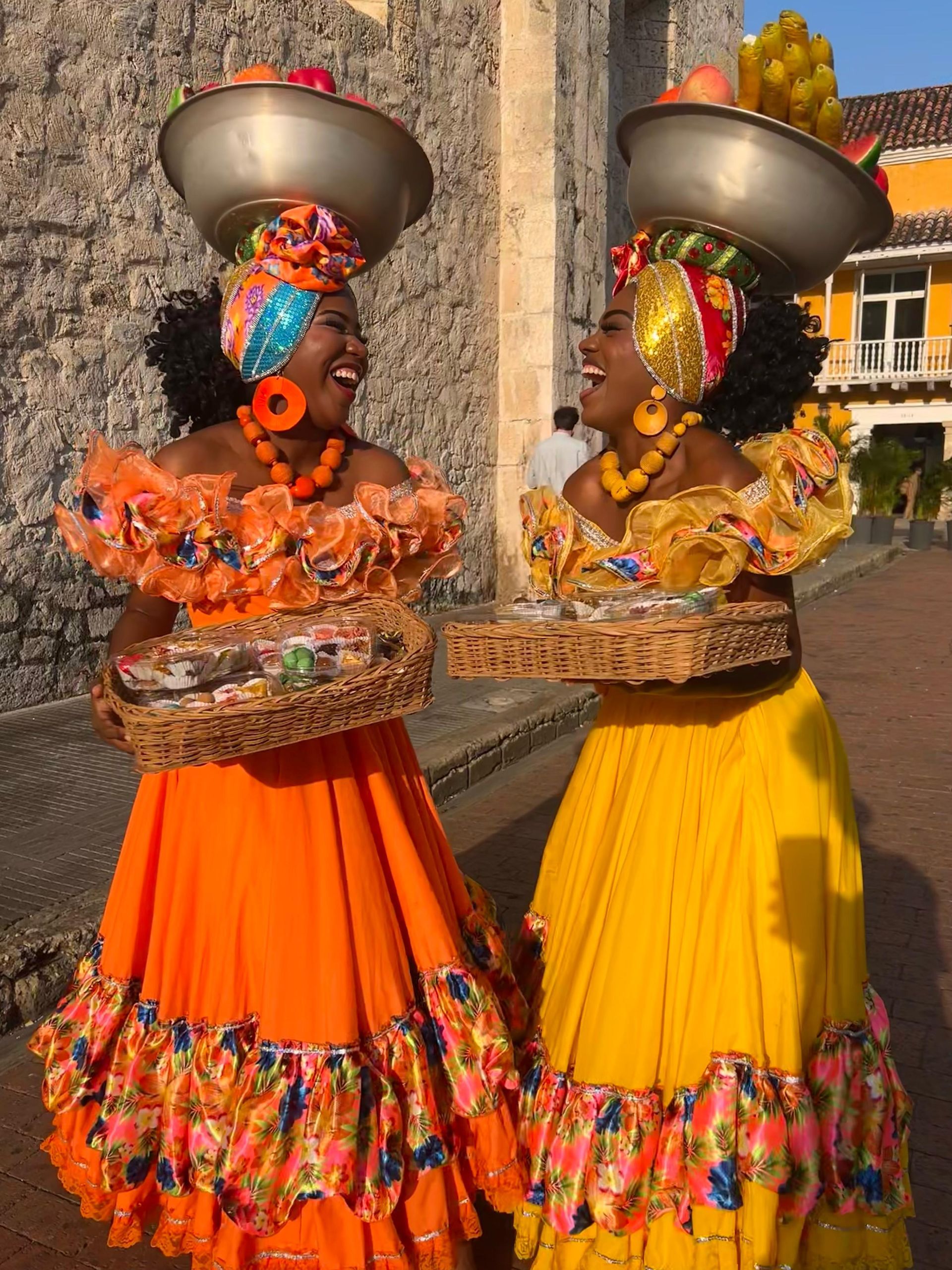 Dos mujeres con vestidos y sombreros brillantes y coloridos balanceando cuencos, riendo en una escena callejera soleada.