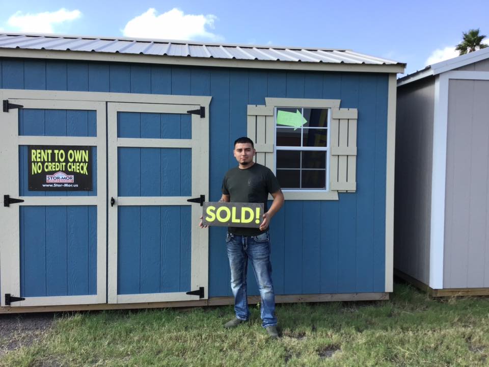 Happy Satisfied Smiling Customer, Domingo, standing in front of his new blue cottage shed with white trim, metal rood, decorative shutters holding a sold sign after he bought his building from Stor-Mor Portable Buildings.