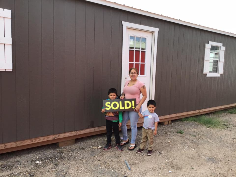Happy Satisfied Smiling Customer, Berenice, standing in front of her new Stor-Mor Portable Building utility building for backyard in brown with white trim holding a sold sign with her small son children after buying her building from Stor-Mor Portable Buildings - South Tex Portable Buildings.