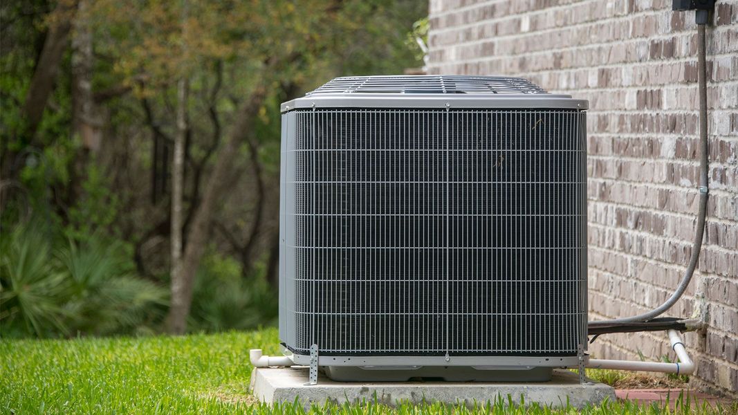 A gray central air conditioning unit sits on a concrete pad next to a brick exterior wall near a grassy lawn and trees.
