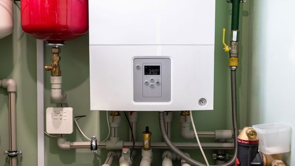 A white wall-mounted boiler with a digital display, connected to pipes and a red expansion tank against a green wall.