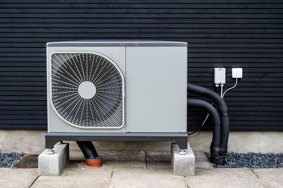 A gray outdoor heat pump unit mounted on concrete blocks against a dark horizontal-slatted wall.
