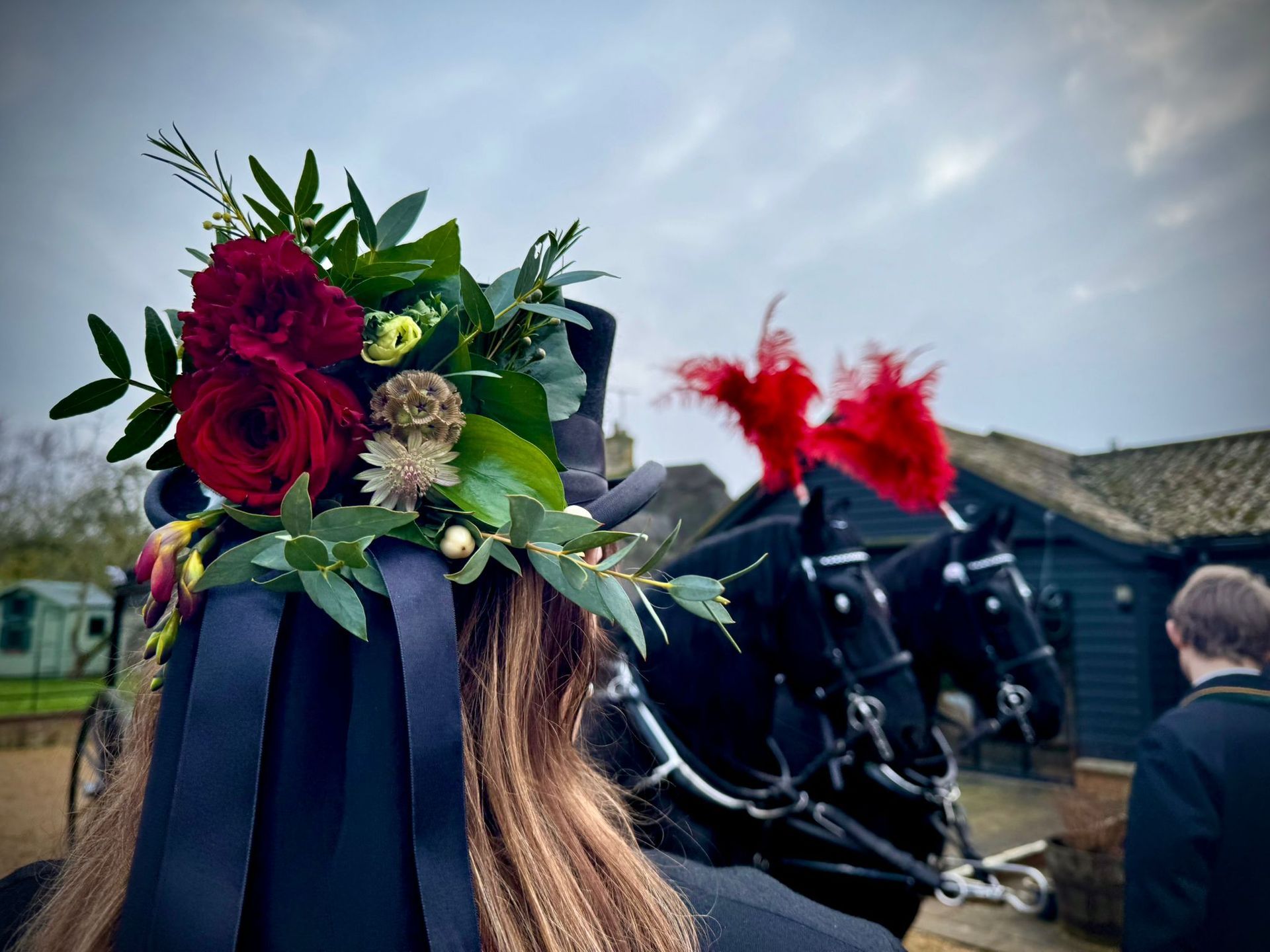 Woman with floral hat stands before a black horse-drawn carriage with red plumes.
