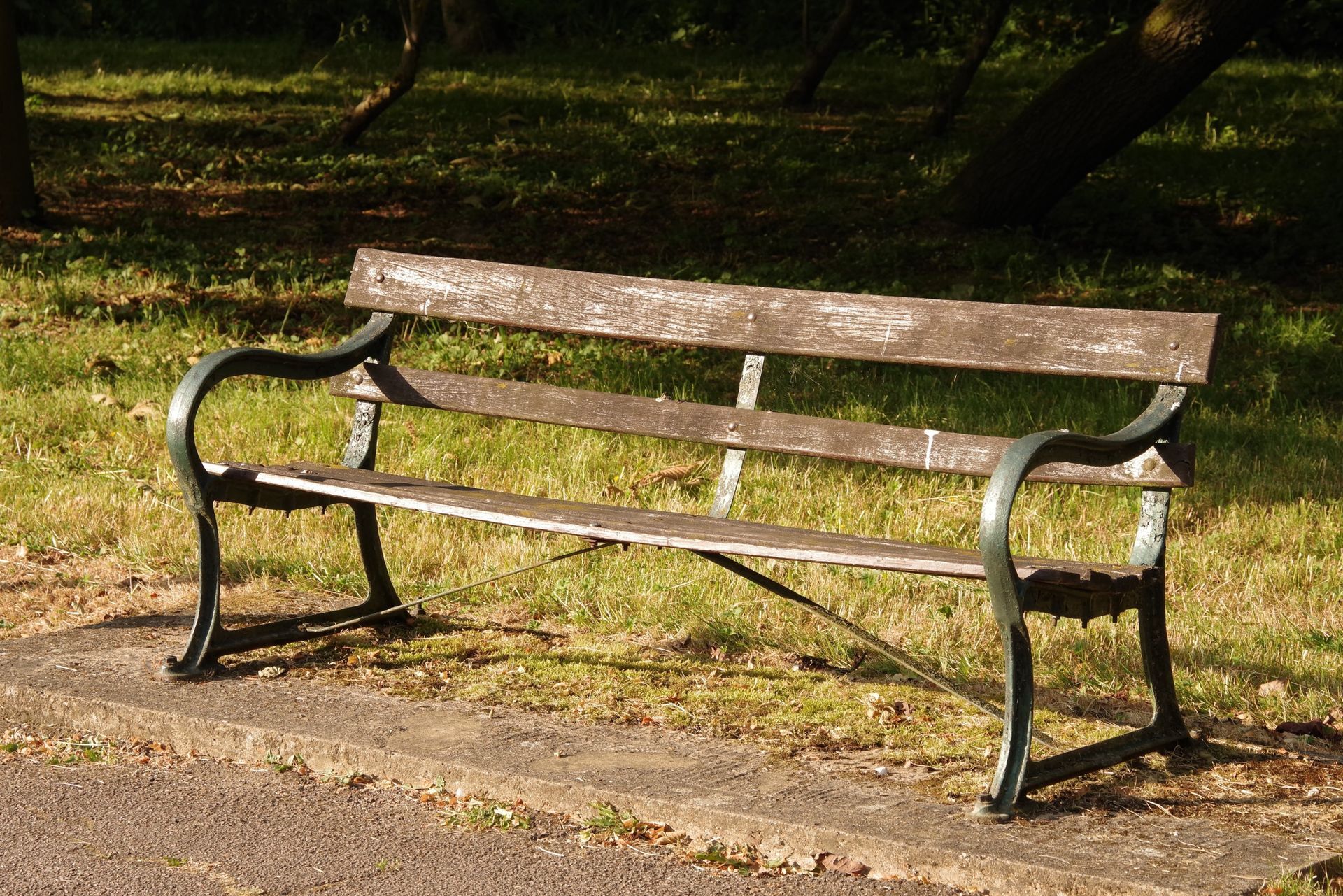 Park bench with worn wooden planks and black metal frame, set on a paved path with grass and trees.