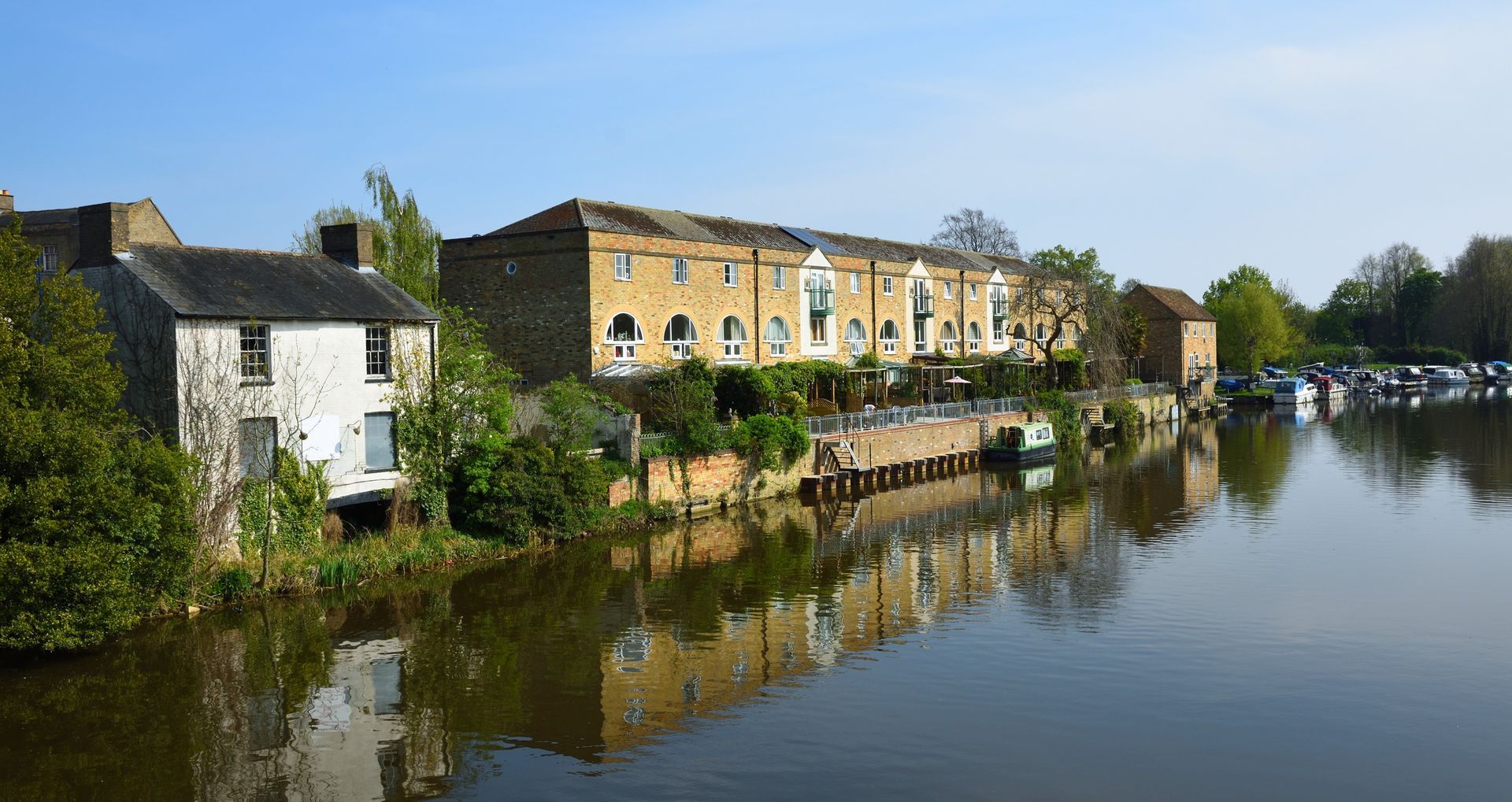 Row of waterfront buildings reflecting in calm water. Trees and a clear blue sky.