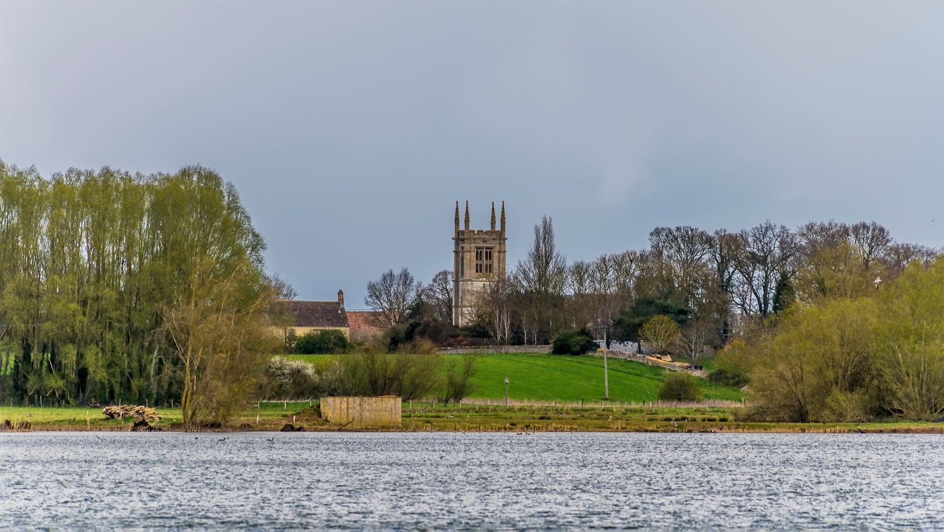A church tower rises above trees and a green hillside, viewed from across a lake under a cloudy sky.