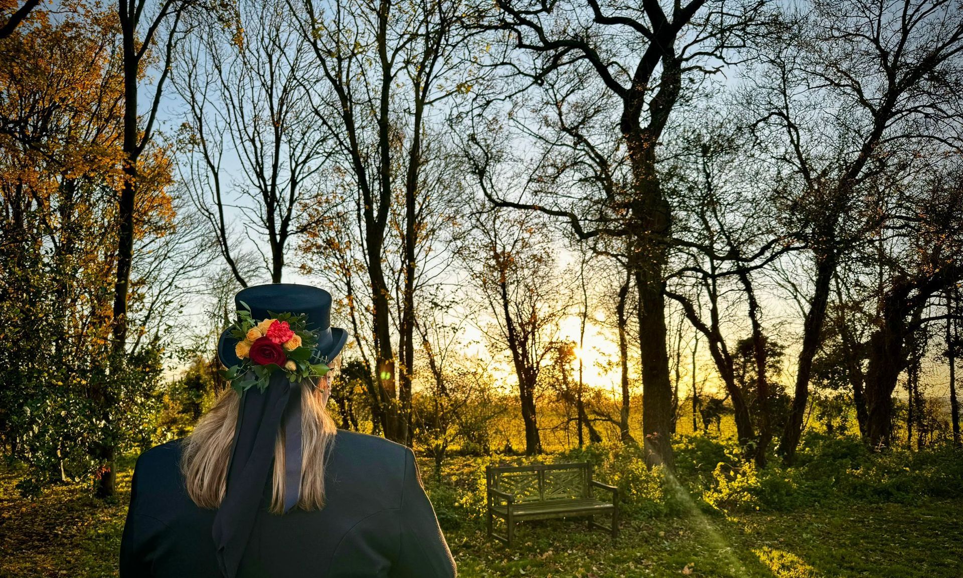 Person in black coat and top hat with floral crown, facing forest at sunset.