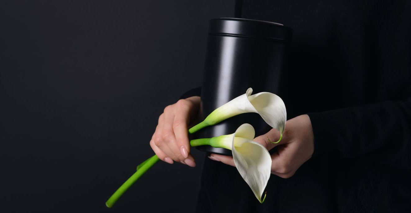 Woman in black holds a black urn and two white calla lilies against a dark background.
