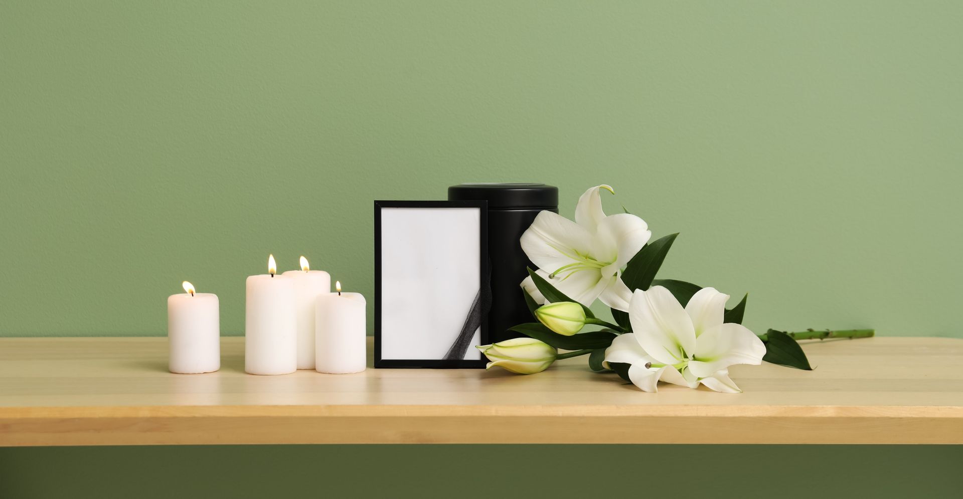 Candles, lilies, an urn, and a framed photo on a wooden surface against a green wall, suggesting a memorial.
