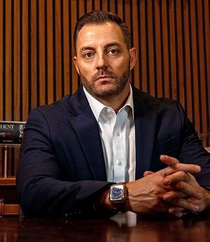 Man in a navy suit seated at a table, hands clasped, against a wood-paneled wall.