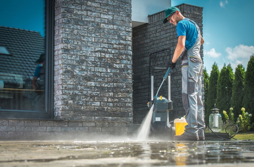 A Man is Using a High Pressure Washer to Clean a Driveway in Front of a Brick Building — Reach 'N' Clean in Ballina, NSW