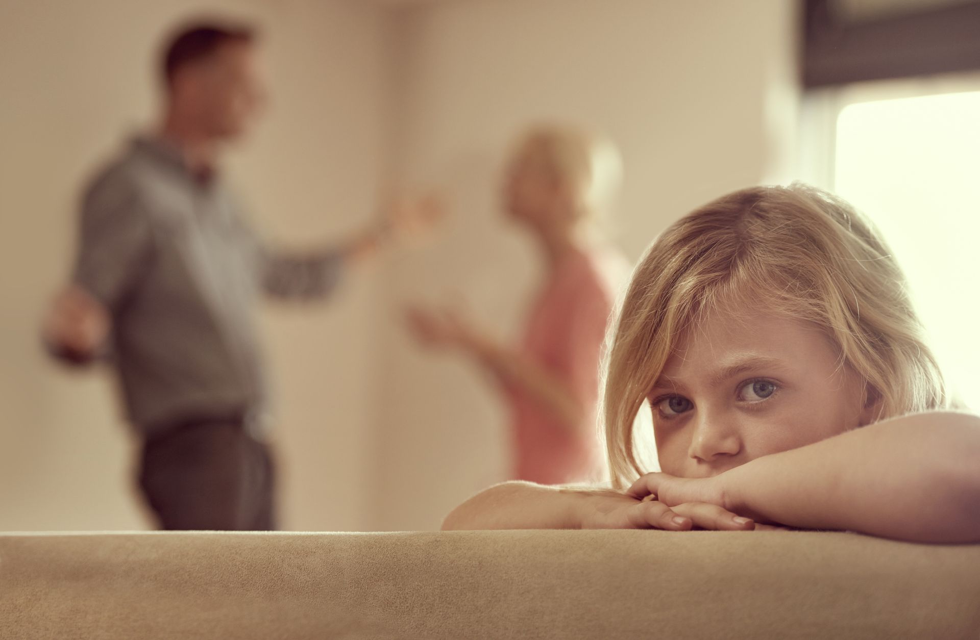 A little girl is sitting on a couch while her parents argue in the background.