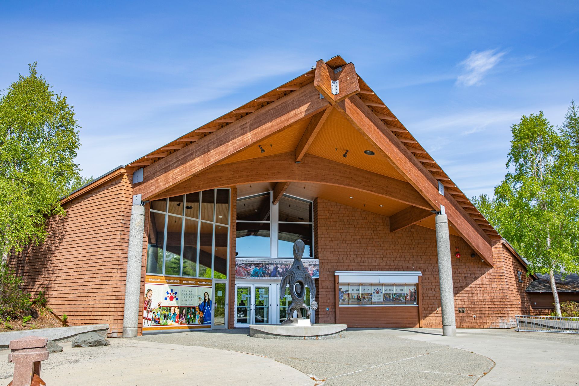 Alaska Native Heritage Center with a sloped roof sits by a road with a crosswalk under a clear, bright blue sky.