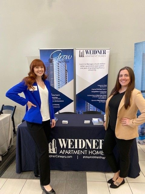 Two smiling professionals stand in front of a blue Weidner Apartment Homes booth with branded banners at an event.