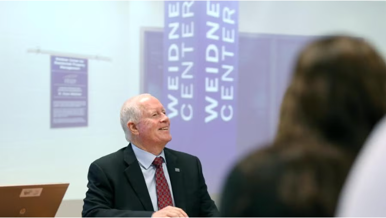 A person in a suit and tie smiles while looking upward in front of a purple Weidner Center sign in a brightly lit room.