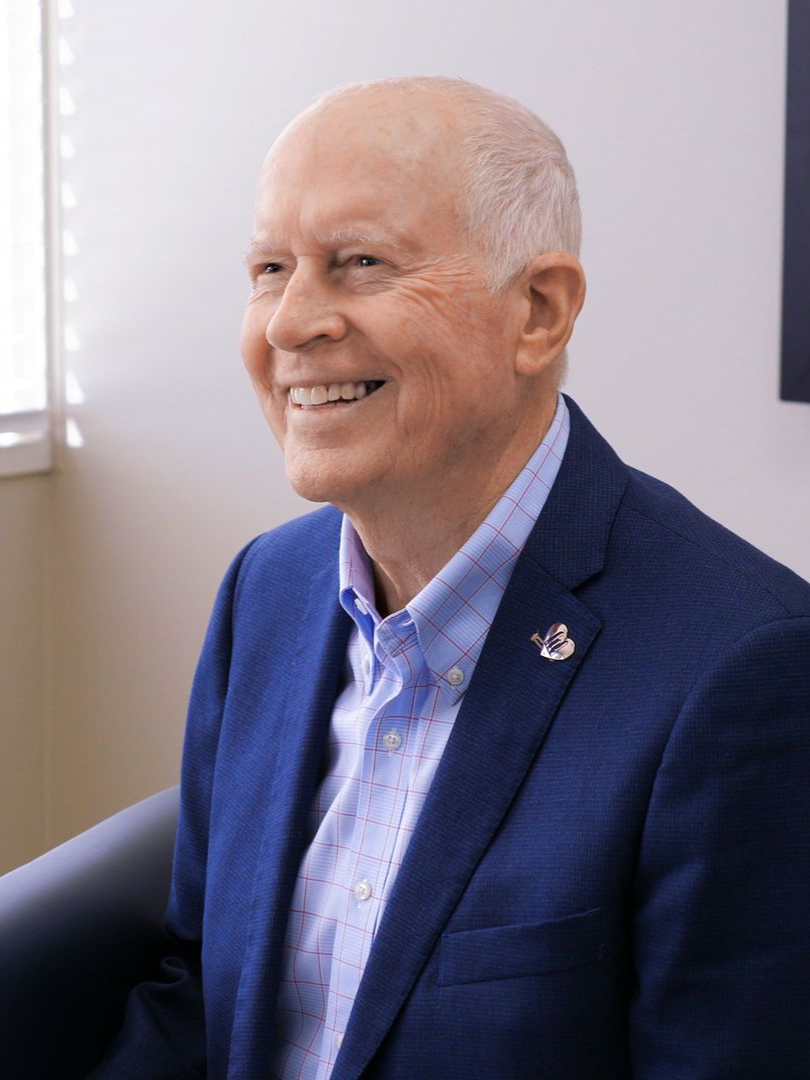 Dean Weidner in a blue blazer and light blue patterned shirt smiling and looking to their right in a bright, indoor setting.