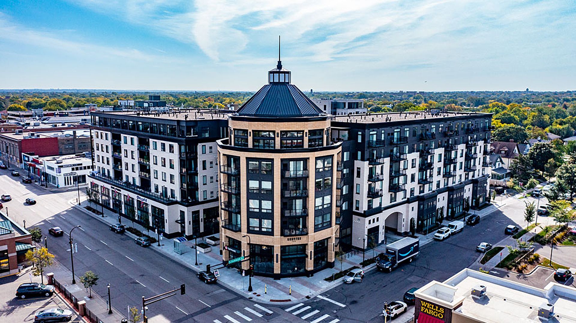 An aerial view of a modern, multi-story apartment building with a distinctive corner turret on a sunny city street.