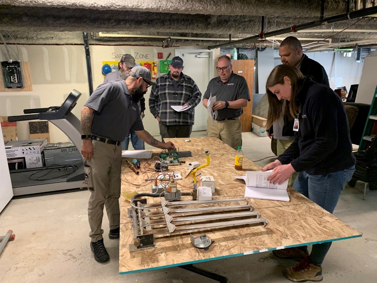 Five people gather around a large table in a workshop, examining mechanical parts and papers for a technical training.