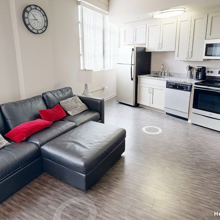 Modern apartment interior with black leather sofa, kitchen appliances, and hardwood floors.