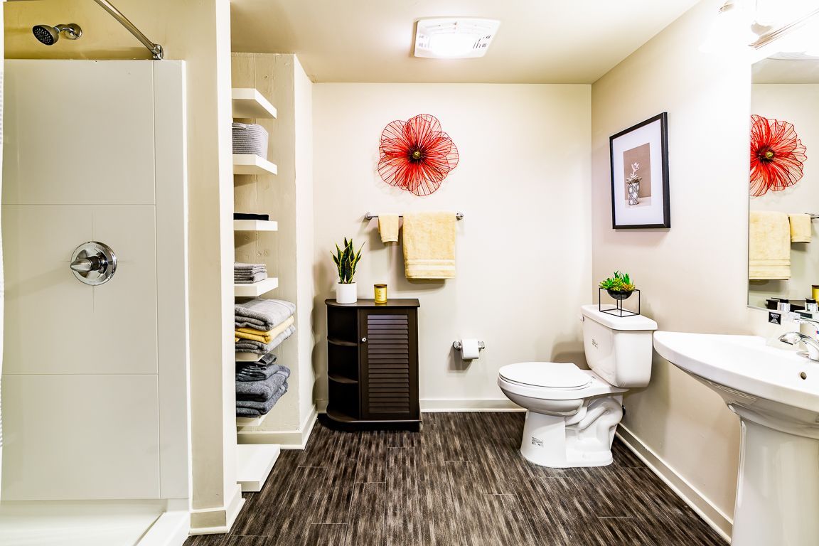 Bathroom with shower, toilet, sink, shelves, and dark wood-look flooring. Red floral art adorns the wall.