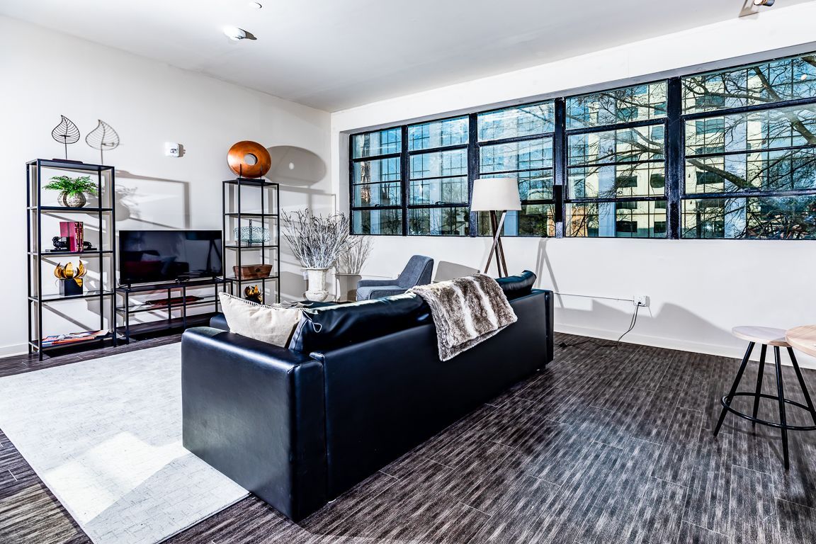 Modern living room with black leather couch, large windows, and shelving.