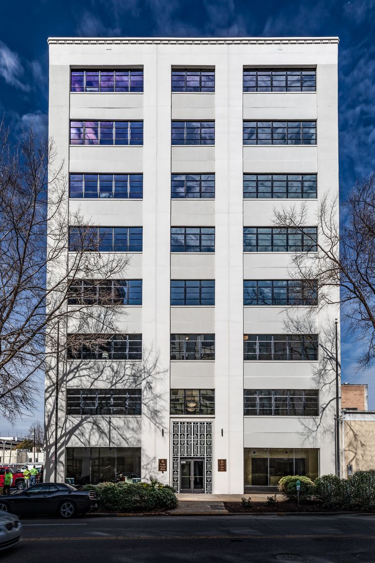 White, multi-story office building with rows of windows against a blue sky. Trees and cars are in the foreground.