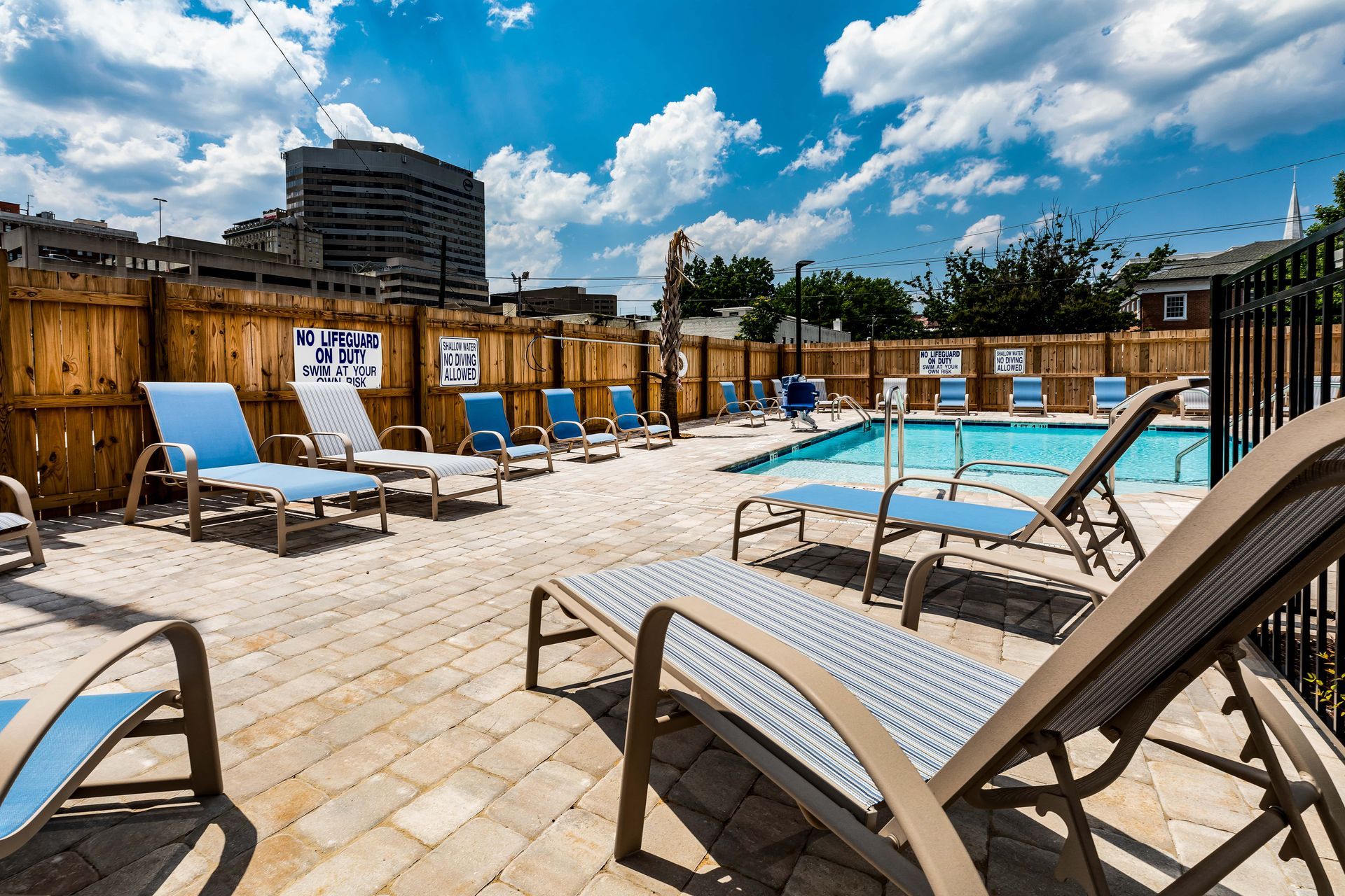 A pool area with blue lounge chairs, a wooden fence, and a building in the distance under a partly cloudy sky.