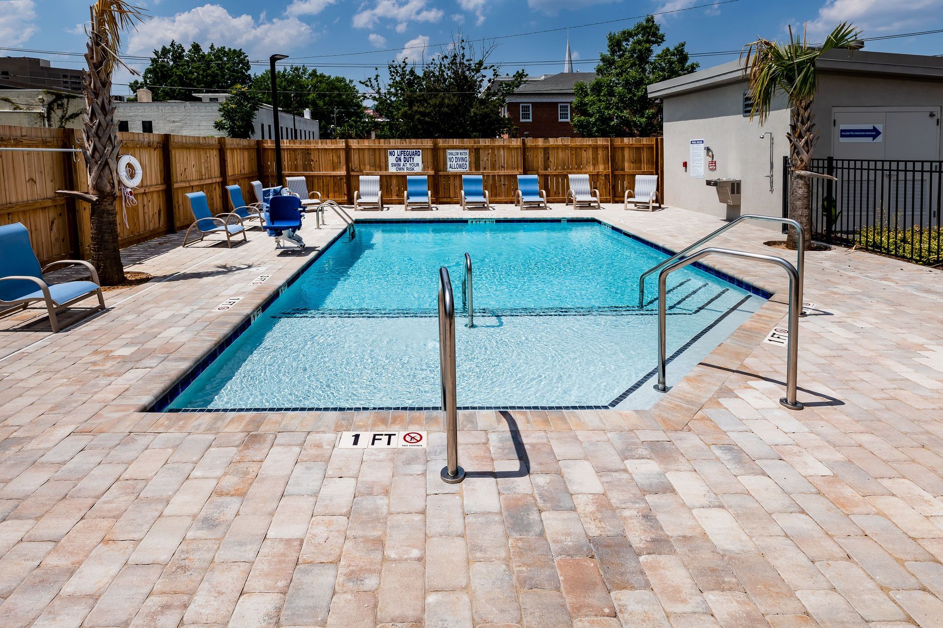 A rectangular swimming pool with lounge chairs, surrounded by a brick patio, wooden fence, and palm trees.