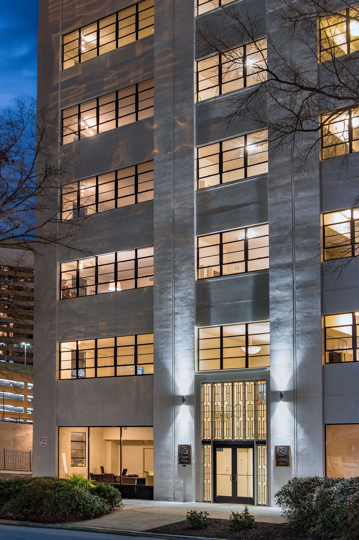 A tall, light-colored office building at dusk, with lit windows, a glass entrance, and illuminated details.