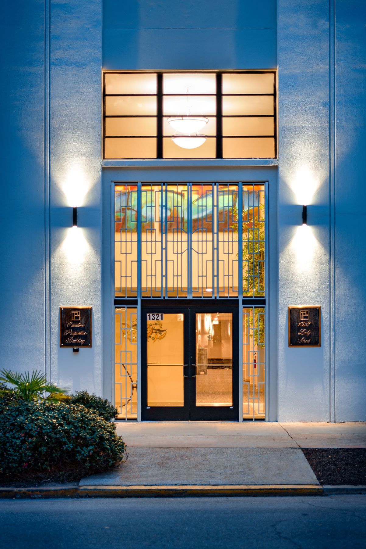 Facade with glass doors, lit at dusk.  A light shines above the doors and two wall sconces light the building.