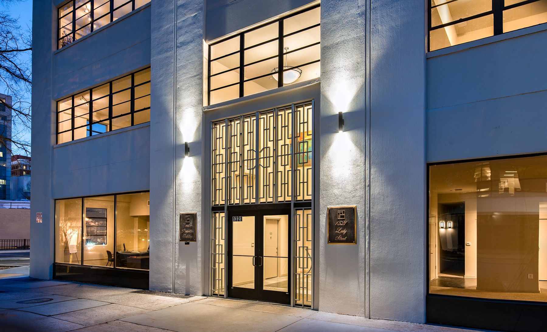Exterior of modern building with black framed windows and entry doors, lit by exterior wall sconces.