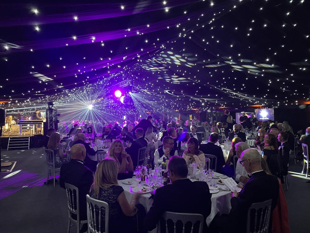 A group of people are sitting at tables in a large room with purple lights.
