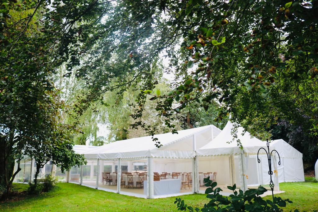 A large white tent is sitting in the middle of a grassy field surrounded by trees.