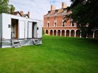 A white trailer is parked in a grassy field in front of a large brick building.