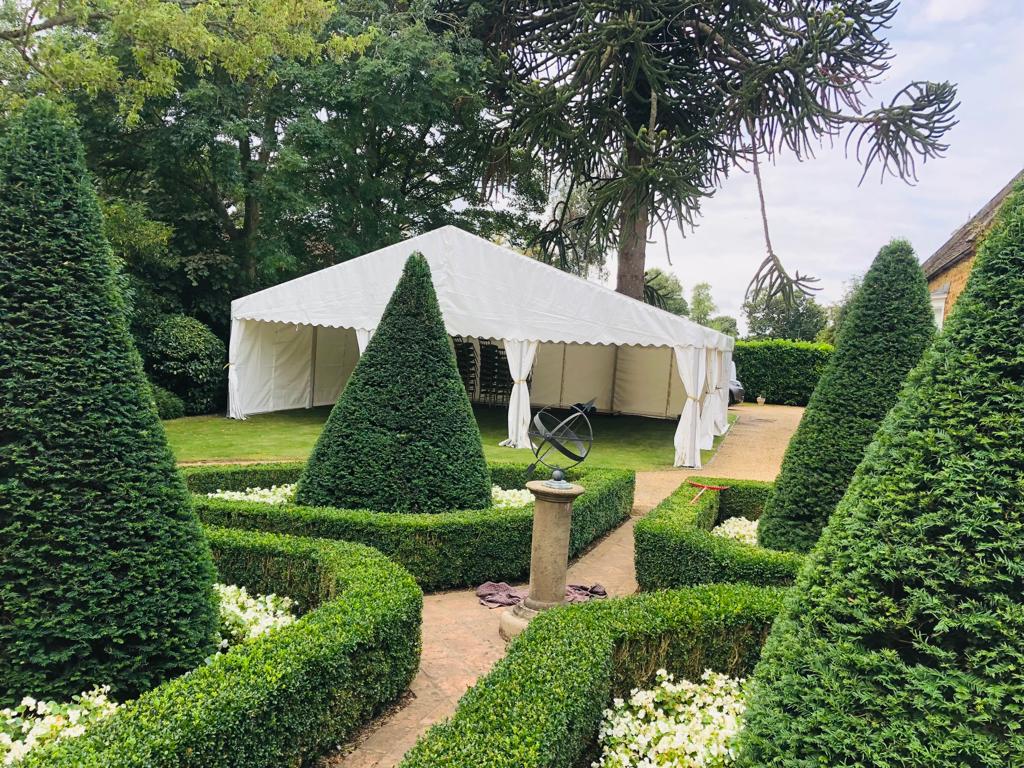 A white tent is sitting in the middle of a garden surrounded by bushes.