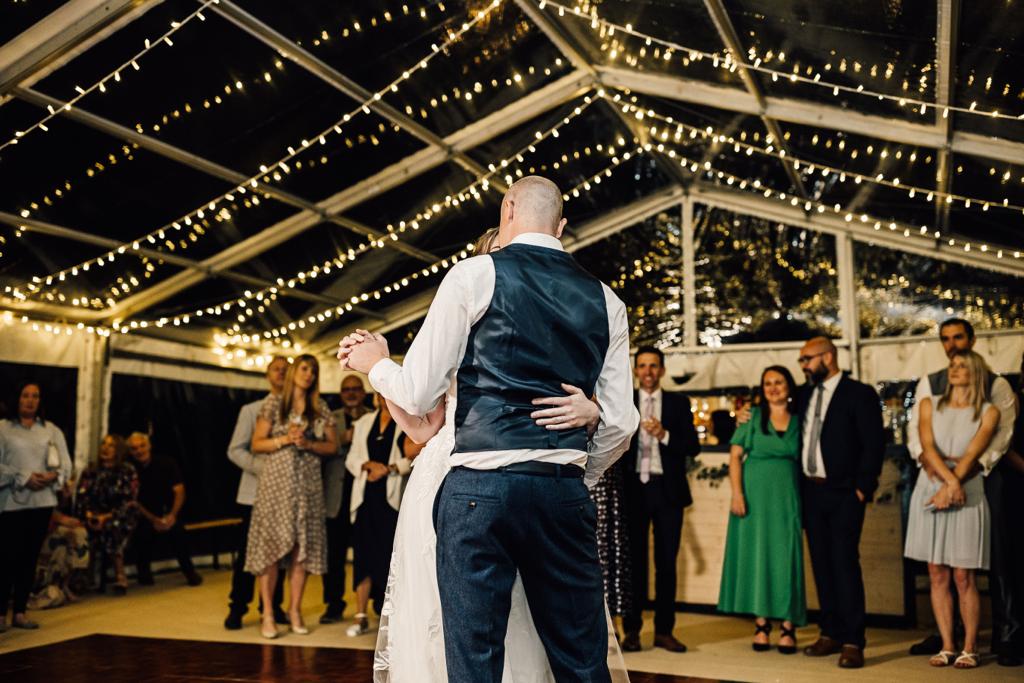 A bride and groom are dancing under a clear tent at their wedding reception.