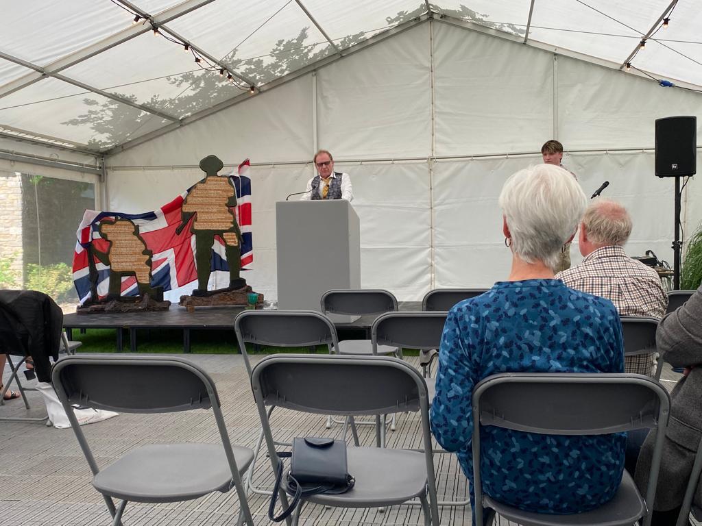 A group of people are sitting in chairs under a tent watching a man giving a speech.