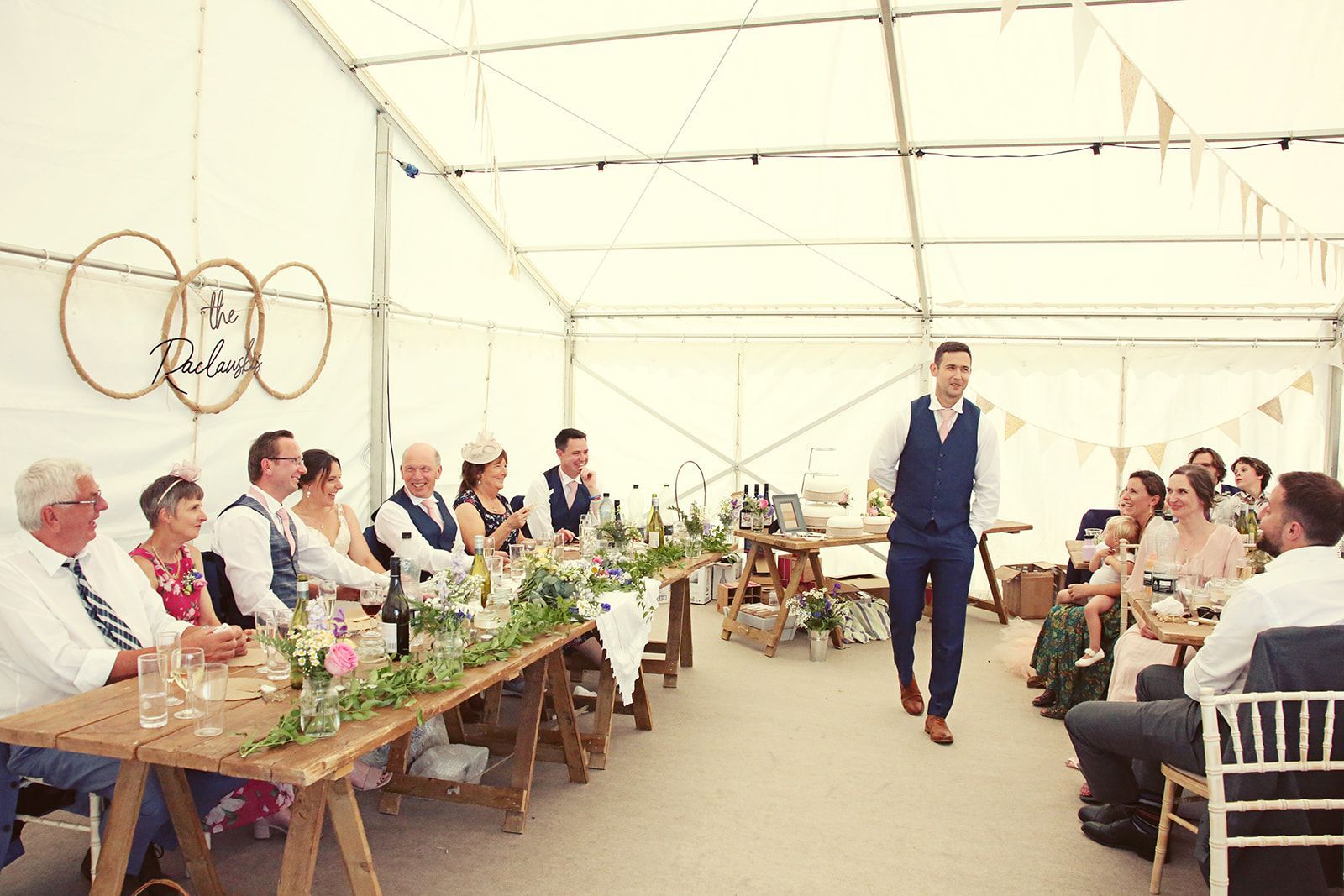 A man is standing in front of a long table at a wedding reception.