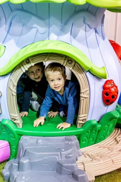 Two young children smiling and looking out of a green and tan playhouse with a blue interior.