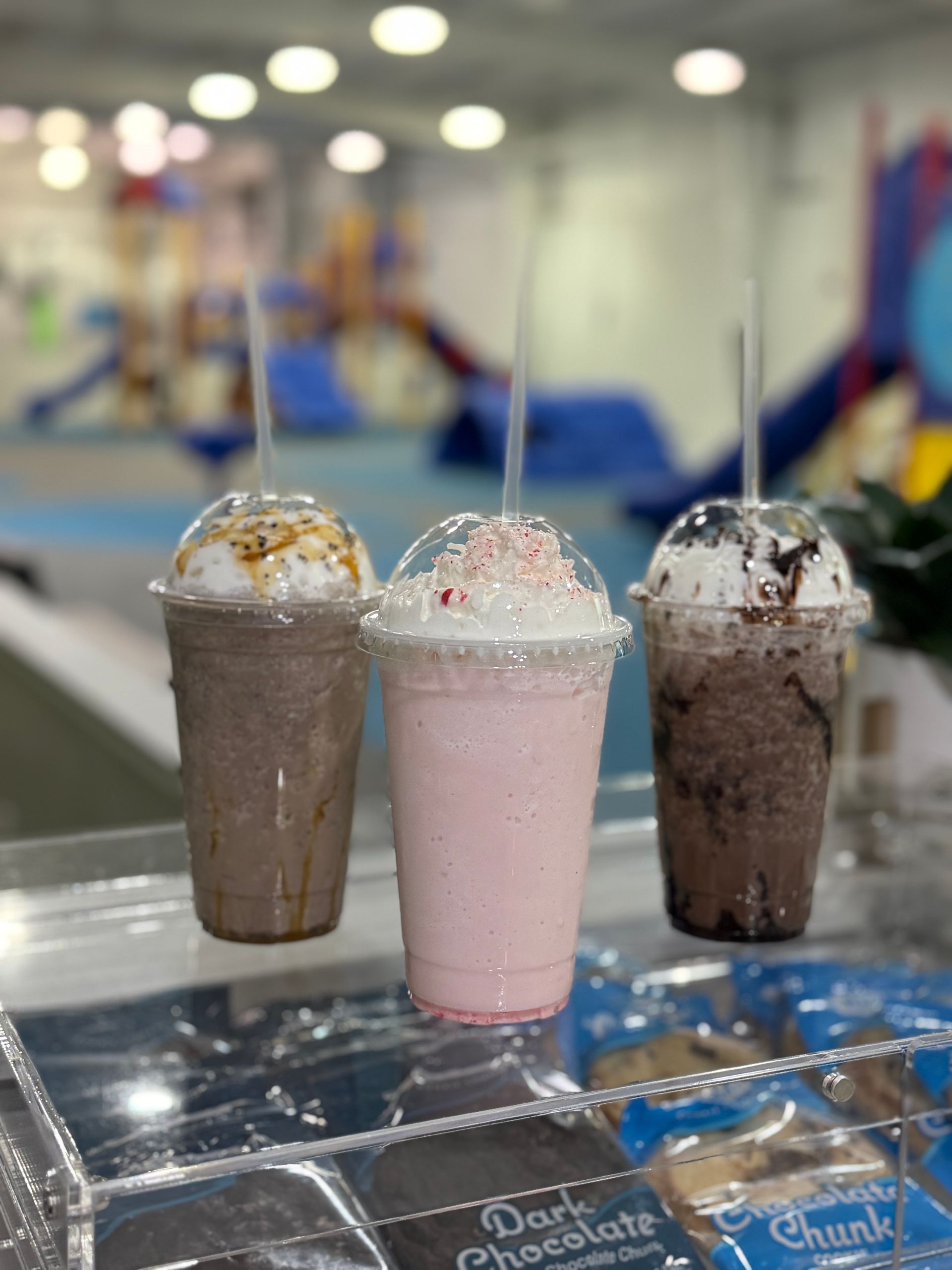 Three milkshakes with toppings, in plastic cups, on a counter with blurred playground background.