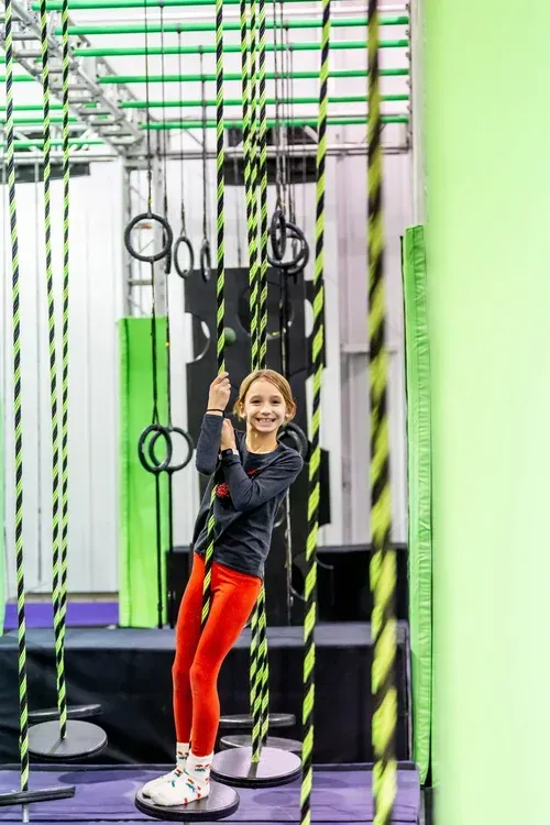 Girl swinging on a rope obstacle course, smiling, wearing red pants and black top.