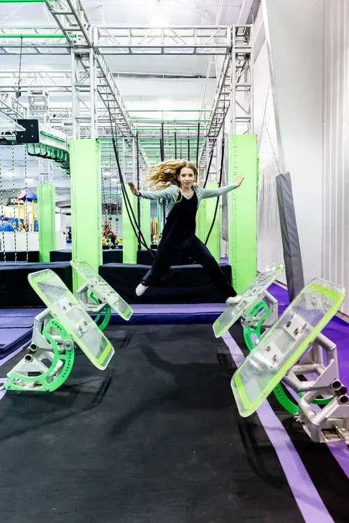 Woman jumping between obstacle platforms at an indoor ninja warrior gym. Green, purple, and black colors are present.