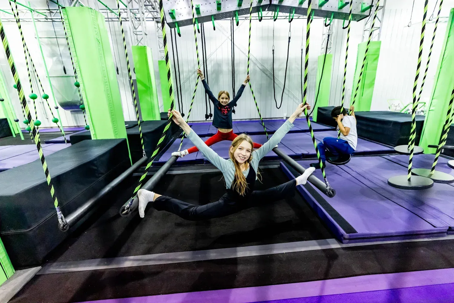 Three people in active poses on trampolines, in a recreational indoor facility with ropes and climbing structures.