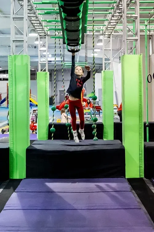 Girl hanging from a bar, crossing an obstacle course. Indoor gym with green and black structures.