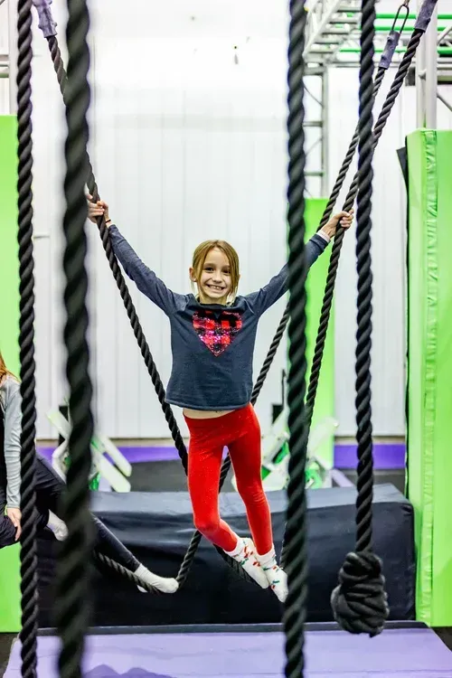 Girl smiles as she swings on ropes in an indoor obstacle course; red pants, gray shirt.