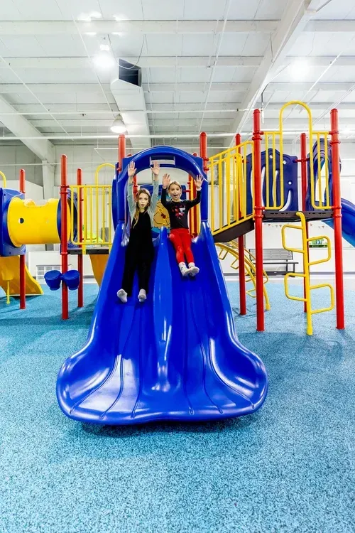 Two children on a blue slide at an indoor playground, waving with smiles.