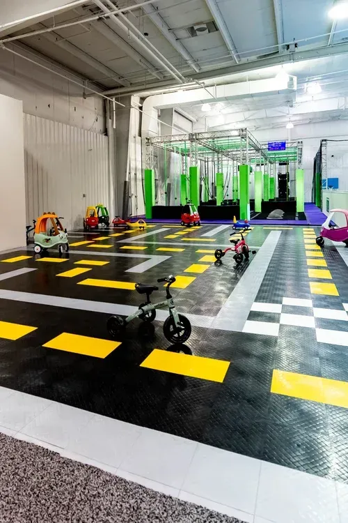 Indoor play space with colorful toy cars on a black, yellow, and white road-like floor; an obstacle course is visible in the background.