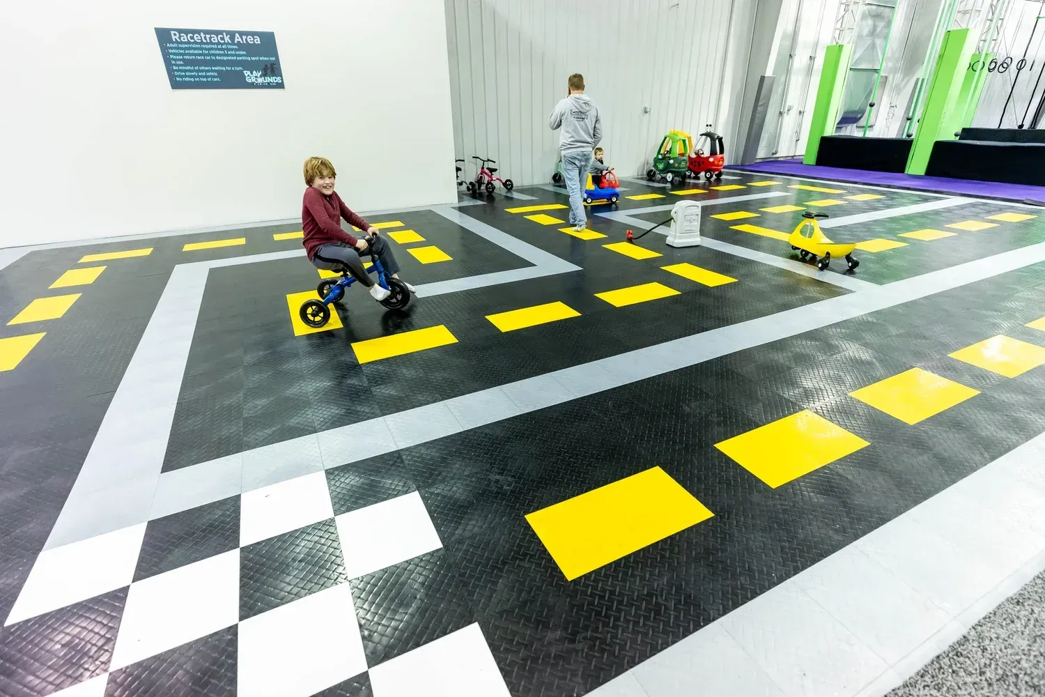 Child rides a trike on a road-themed floor with others. An adult walks nearby. Indoor setting with gray, black, and yellow.