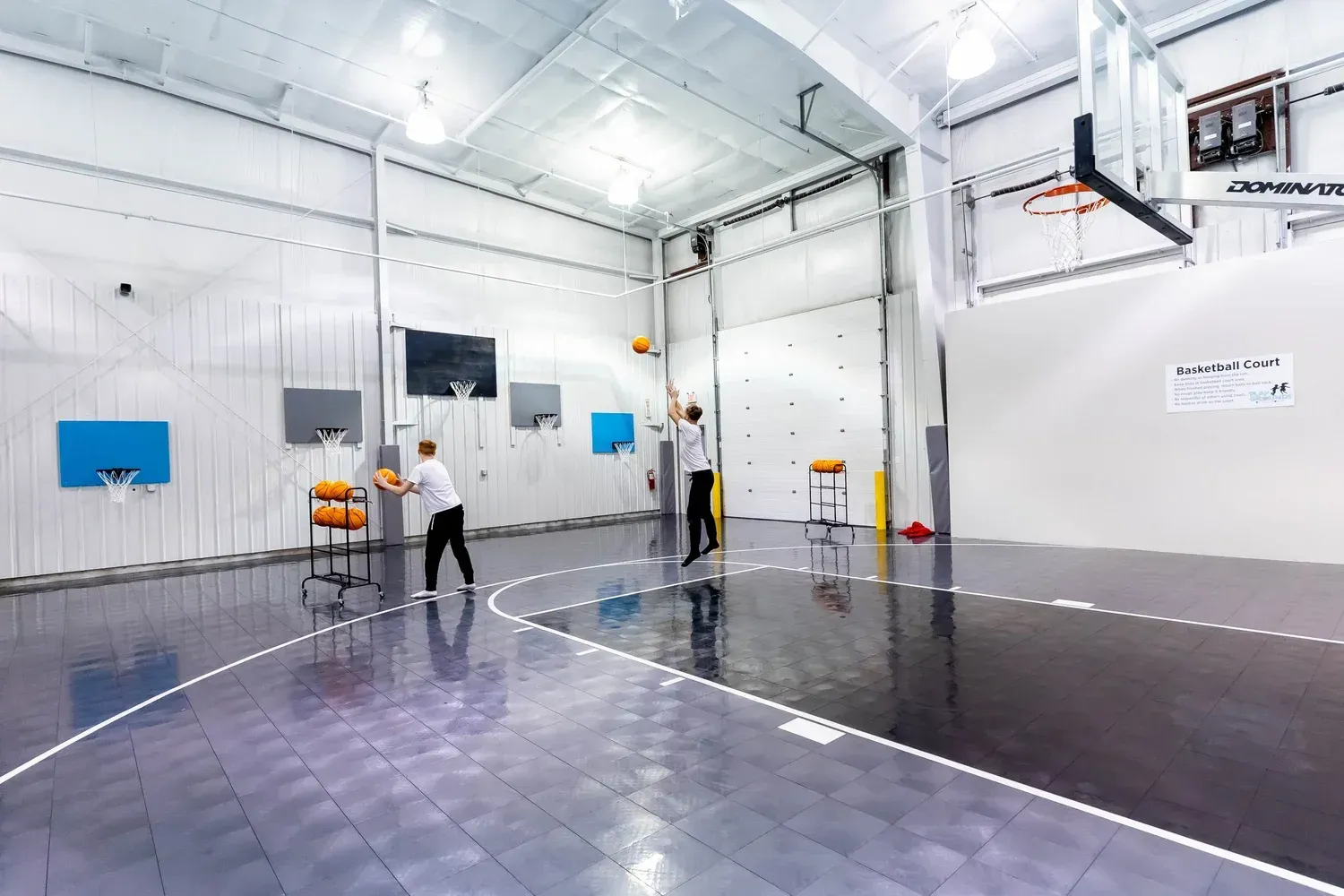 Two people playing basketball in an indoor court. One throws a ball towards the hoop. Bright, spacious facility.